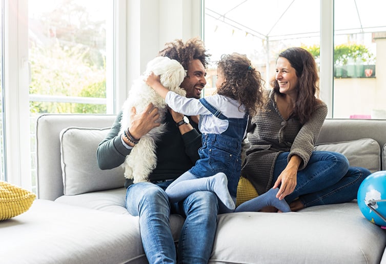 Man, woman, child, and dog sitting on a gray couch together laughing