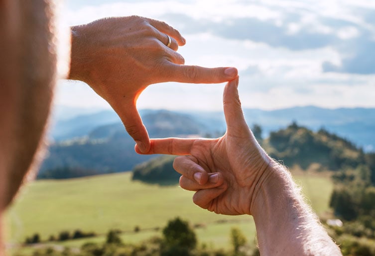 Close up of someone's hands. They're forming a form with their thumbs and pointer fingers, looking out at the sky in the distance