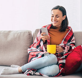 Woman sitting on couch, smiling at her phone. She's wearing a red plaid blanket