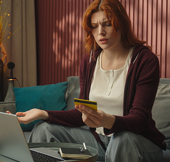 Person seated on a sofa in a living room, holding a yellow card while looking at a laptop on a nearby table. A notebook and pen rest beside the laptop. The room features teal cushions and warm lighting against a paneled wall.