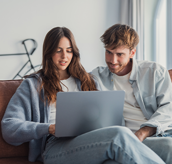 Two people sitting together on a couch, looking at a laptop screen. A bicycle and large windows are visible in the background of a bright indoor setting.