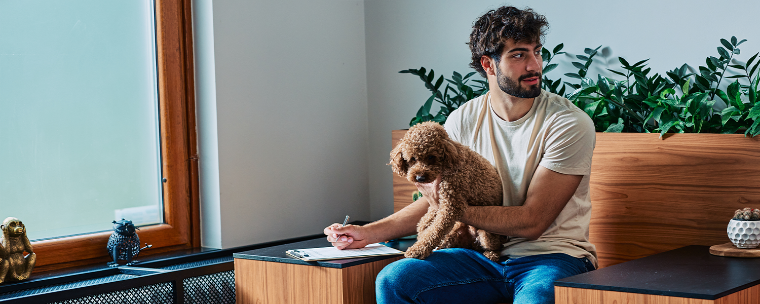 Person seated on a wooden bench with a small dog on their lap, writing on a clipboard in a modern indoor space with plants in the background.
