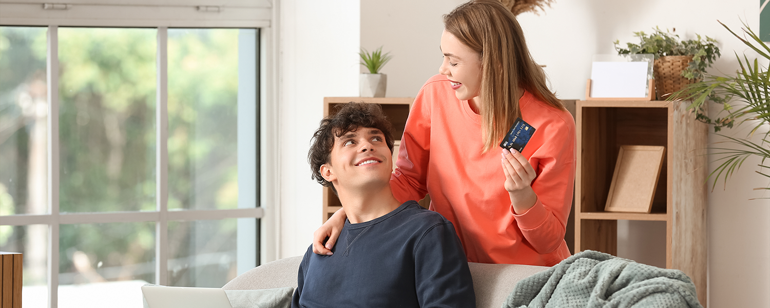 Two people in a living room beside a laptop. One person sits in an armchair using the laptop while the other stands nearby holding a credit card. A blanket, bookshelf, and large window are visible in the background.