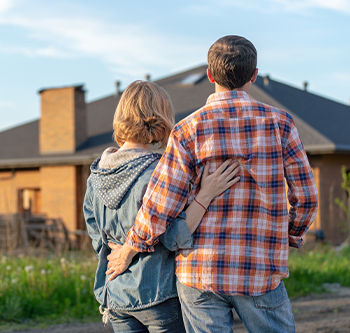 Two people standing outdoors with their backs to the camera, looking toward a house. One person has an arm around the other, with a grassy area and residential neighborhood visible in the background.