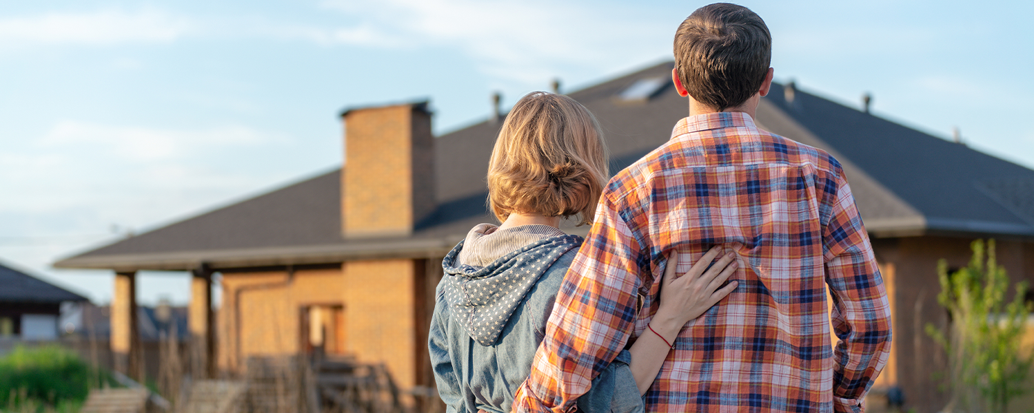 Two people standing outdoors with their backs to the camera, looking toward a house. One person has an arm around the other, with a grassy area and residential neighborhood visible in the background.