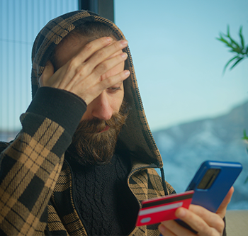 Person wearing a plaid hooded sweater sits indoors holding a smartphone and a credit card, with one hand covering part of the forehead. A plant and window are visible in the background.