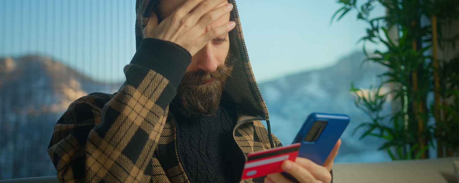 Person wearing a plaid hooded sweater sits indoors holding a smartphone and a credit card, with one hand covering part of the forehead. A plant and window are visible in the background.