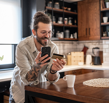 Person standing at a kitchen island, holding a smartphone and credit card while looking at the phone, with a coffee cup and kitchen cabinets visible in the background.