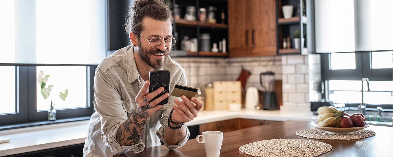 Person standing at a kitchen island, holding a smartphone and credit card while looking at the phone, with a coffee cup and kitchen cabinets visible in the background.