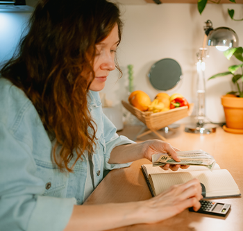 Person seated at a kitchen counter using a calculator beside an open notebook and a stack of cash. A bowl of fruit, potted plants, and a desk lamp are visible in the background.