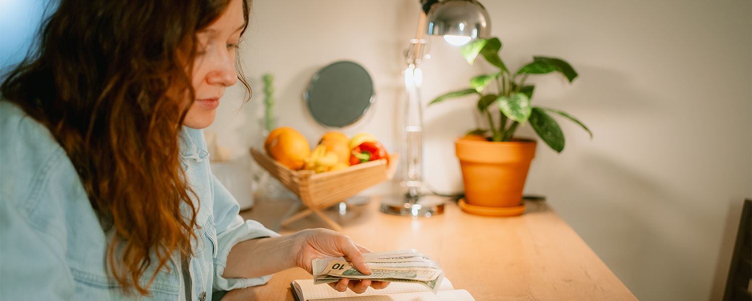 Person seated at a kitchen counter using a calculator beside an open notebook and a stack of cash. A bowl of fruit, potted plants, and a desk lamp are visible in the background.