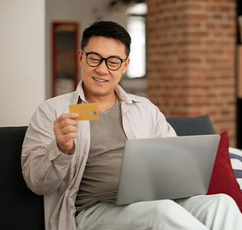 Person sitting on a sofa with a laptop on their lap, holding a credit card while looking toward the computer. Exposed brick walls, a cushion, and a modern indoor living space are visible in the background.