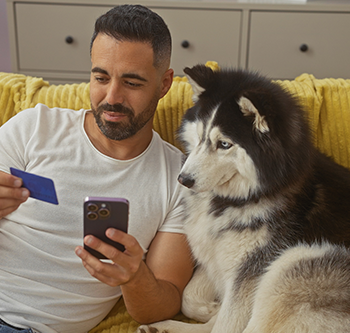 Person sitting on a couch beside a large dog, holding a smartphone and a credit card. A yellow blanket is draped over the couch, and cabinets are visible in the background.