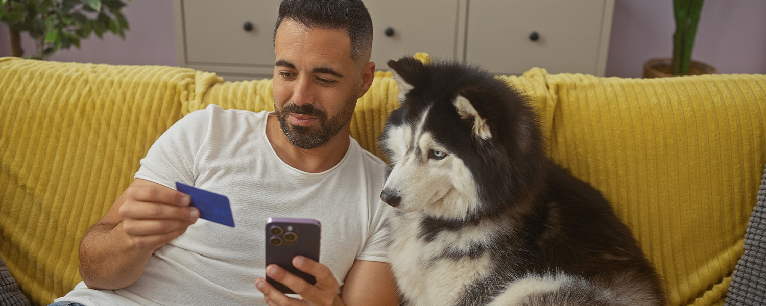 Person sitting on a couch beside a large dog, holding a smartphone and a credit card. A yellow blanket is draped over the couch, and cabinets are visible in the background.