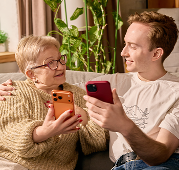 Two people sitting together on a couch, each holding a smartphone and looking at the screens. Houseplants and a window are visible in the background of a well-lit living room.