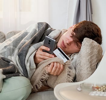 Person lying on a couch wrapped in a gray blanket, holding a smartphone in one hand and a credit card in the other, with a pillow and small table nearby.