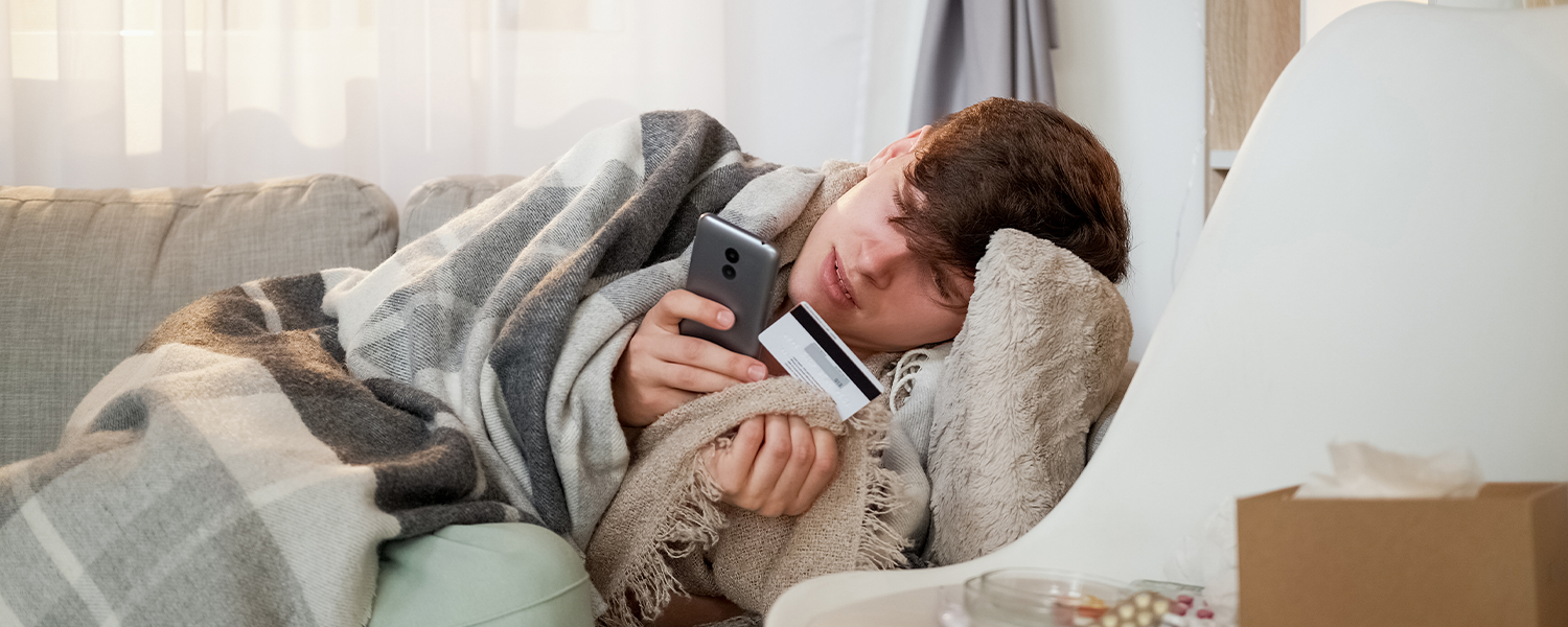 Person lying on a couch wrapped in a gray blanket, holding a smartphone in one hand and a credit card in the other, with a pillow and small table nearby.