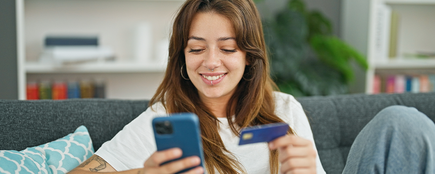 Person sitting on a couch, holding a smartphone in one hand and a credit card in the other while looking at the phone, with decorative pillows and a plant in the background.