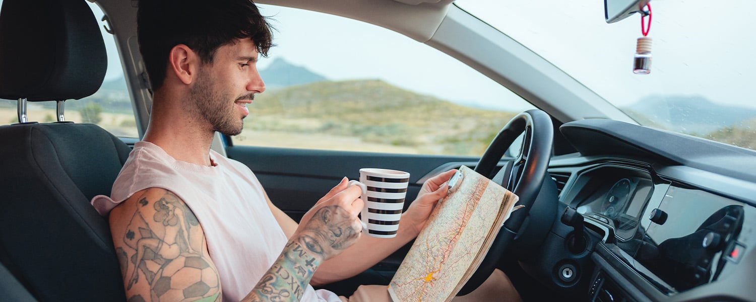Person taking a break inside a parked car, holding a paper map and a coffee mug while planning a road trip route.