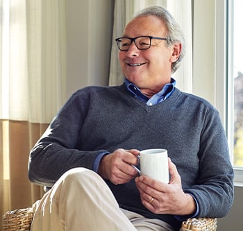Person seated in a sunlit chair near a window, holding a coffee mug and relaxing at home.