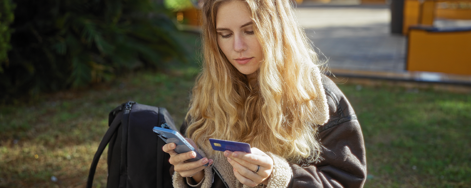 Person sitting outdoors holding a smartphone in one hand and a debit card in the other. A backpack rests nearby, with trees and a walkway visible in the background.