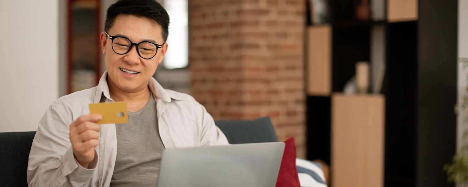 Person sitting on a sofa with a laptop on their lap, holding a credit card while looking toward the computer. Exposed brick walls, a cushion, and a modern indoor living space are visible in the background.