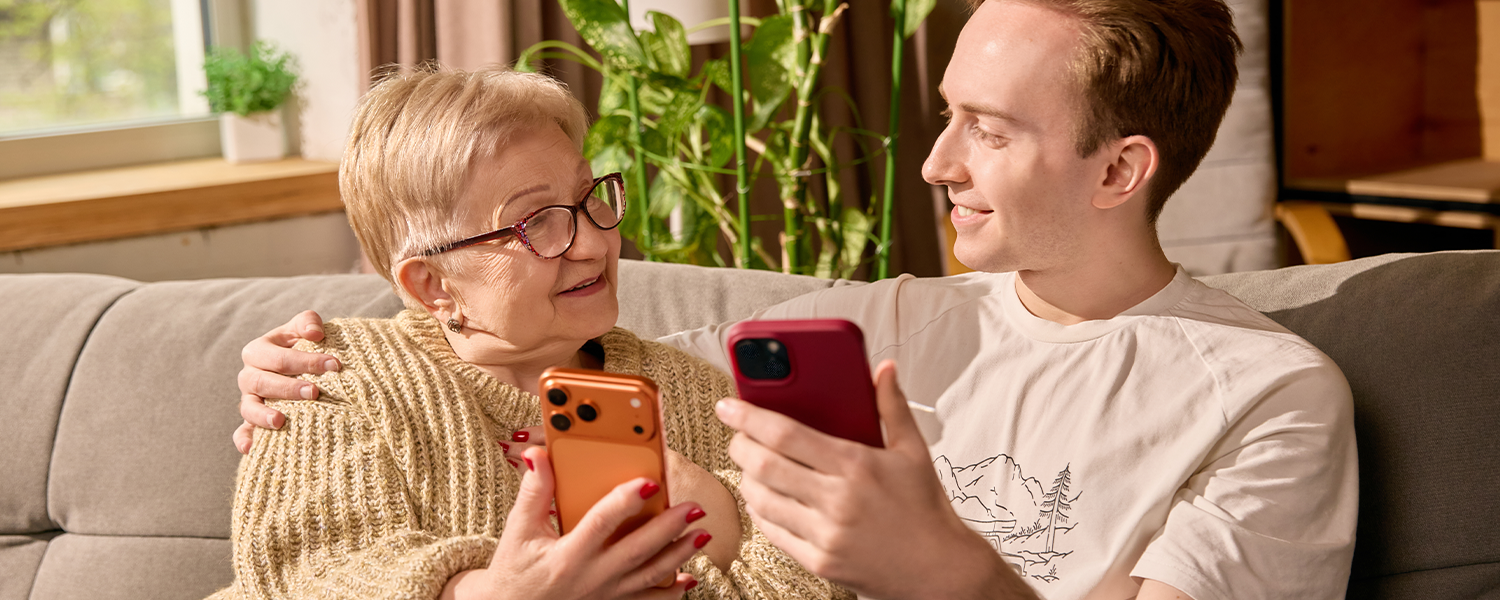 Two people sitting together on a couch, each holding a smartphone and looking at the screens. Houseplants and a window are visible in the background of a well-lit living room.