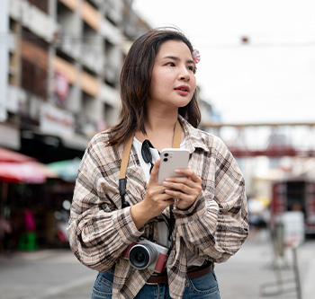 Traveler standing on a busy city street, holding a smartphone with a camera hanging around the neck while navigating an urban destination.