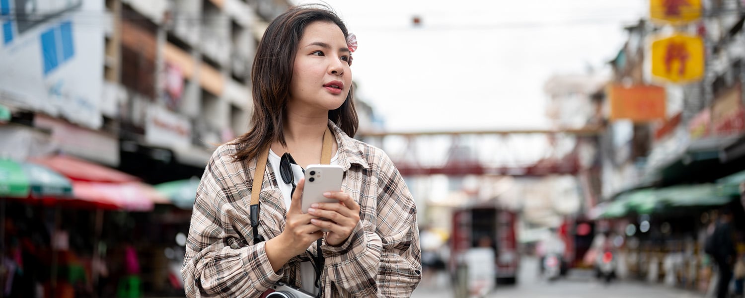 Traveler standing on a busy city street, holding a smartphone with a camera hanging around the neck while navigating an urban destination.