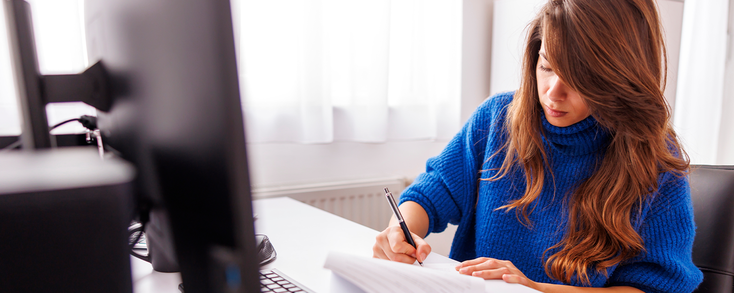 Person seated at a desk writing on paperwork with a pen beside an open laptop. Additional documents are spread across the desk, with a bright window and home office setting in the background.