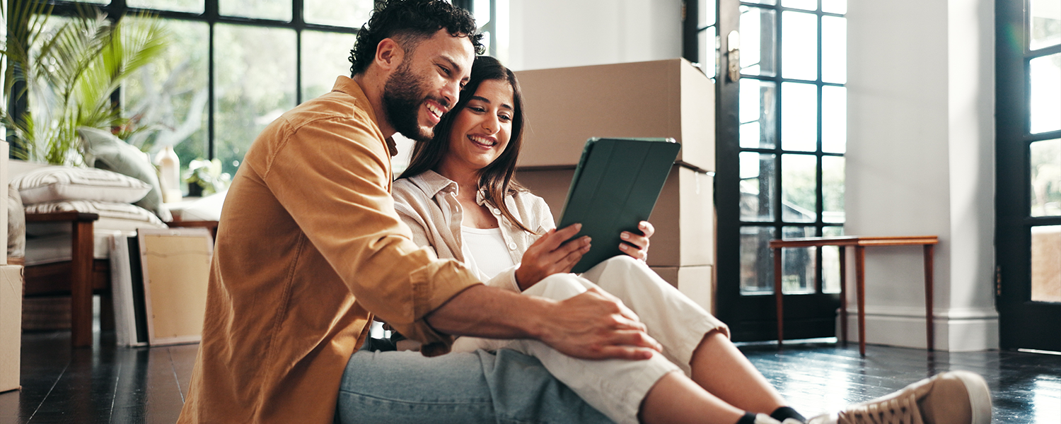 Two people sitting on the floor inside a home with moving boxes nearby, looking at a tablet together. Large windows provide natural light, and the room appears to be in the process of being organized or moved into.