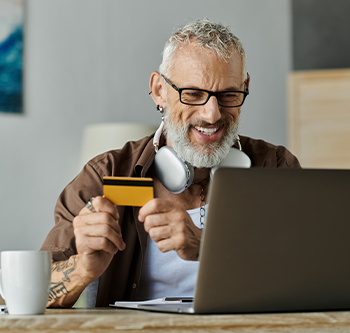 Person seated at a desk using a laptop while holding a credit card, with headphones around the neck, a coffee mug on the desk, and a home office setting in the background.