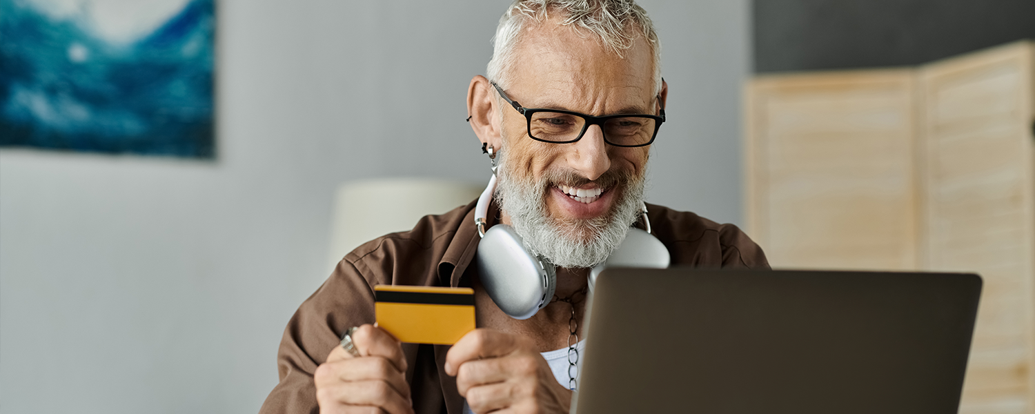 Person seated at a desk using a laptop while holding a credit card, with headphones around the neck, a coffee mug on the desk, and a home office setting in the background.