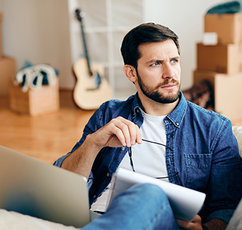 Person seated indoors with a laptop and paperwork, holding a pair of eyeglasses while reviewing documents. Moving boxes and a guitar are visible in the background.