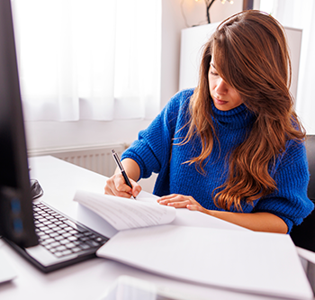 Person seated at a desk writing on paperwork with a pen beside an open laptop. Additional documents are spread across the desk, with a bright window and home office setting in the background.