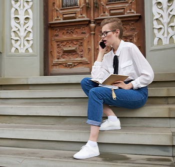 Person sitting on the steps outside a building, talking on a smartphone while holding an open notebook. Decorative wooden doors and patterned architectural details are visible in the background.