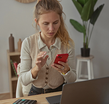 Person seated at a desk, using a red smartphone while a laptop is open nearby. A calculator sits on the desk in the foreground, and a bookshelf and potted plant are visible in the background of the home office setting.