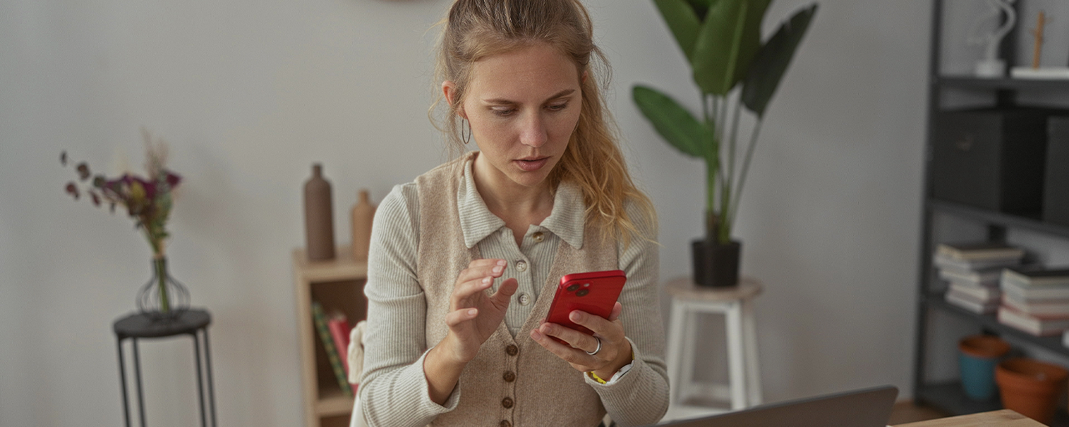 Person seated at a desk, using a red smartphone while a laptop is open nearby. A calculator sits on the desk in the foreground, and a bookshelf and potted plant are visible in the background of the home office setting.