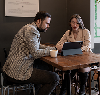 Two people seated at a wooden table reviewing information on a tablet in a meeting space, with a coffee cup on the table and chairs and framed wall decor in the background.