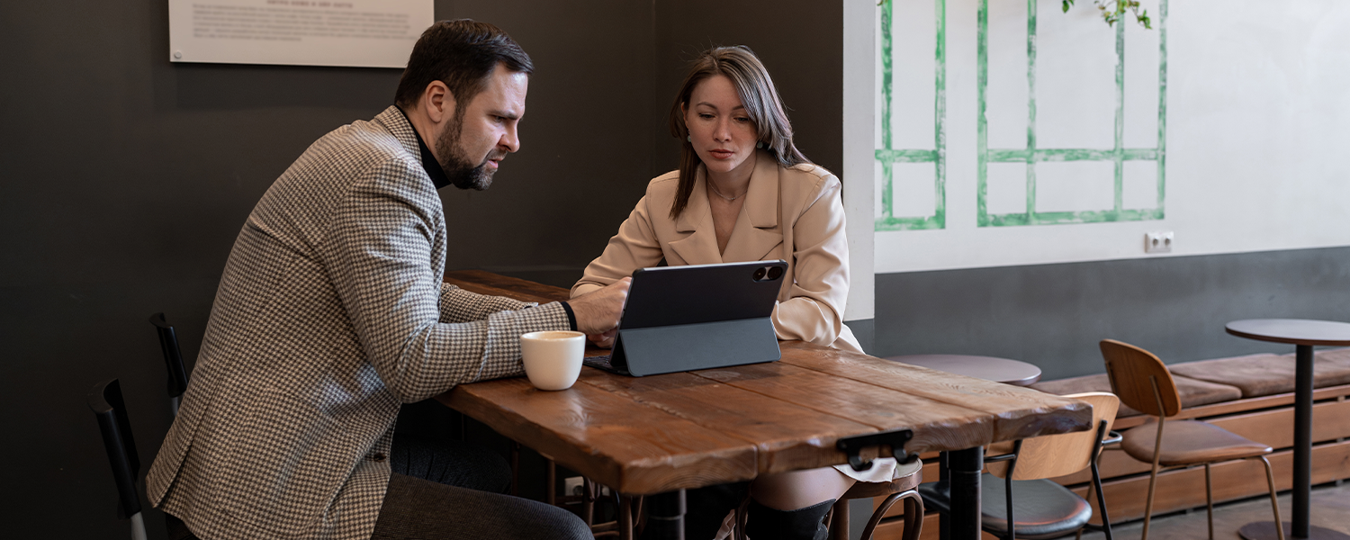 Two people seated at a wooden table reviewing information on a tablet in a meeting space, with a coffee cup on the table and chairs and framed wall decor in the background.