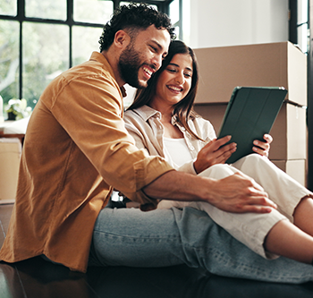 Two people sitting on the floor inside a home with moving boxes nearby, looking at a tablet together. Large windows provide natural light, and the room appears to be in the process of being organized or moved into.
