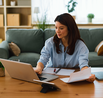Person seated at a wooden coffee table using a laptop while reviewing printed documents and notes. A tablet on a stand sits in the foreground, and a sofa, shelving unit, houseplants, and large windows are visible in the background of a bright living room.
