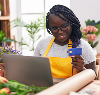 Person wearing a yellow apron sits at a table using a laptop while holding a blue credit card. Flowers, potted plants, and wrapped paper are arranged around the workspace, creating a bright home-based business or creative workspace setting.