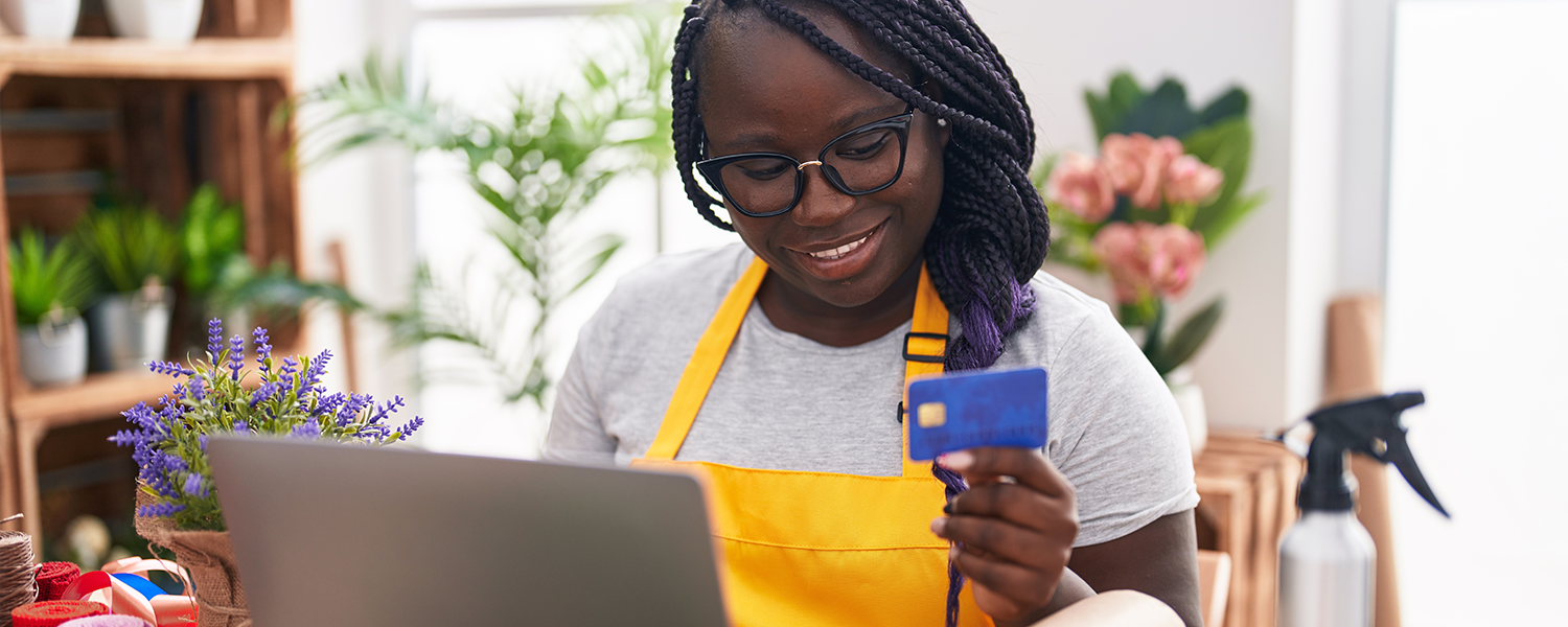 Person wearing a yellow apron sits at a table using a laptop while holding a blue credit card. Flowers, potted plants, and wrapped paper are arranged around the workspace, creating a bright home-based business or creative workspace setting.