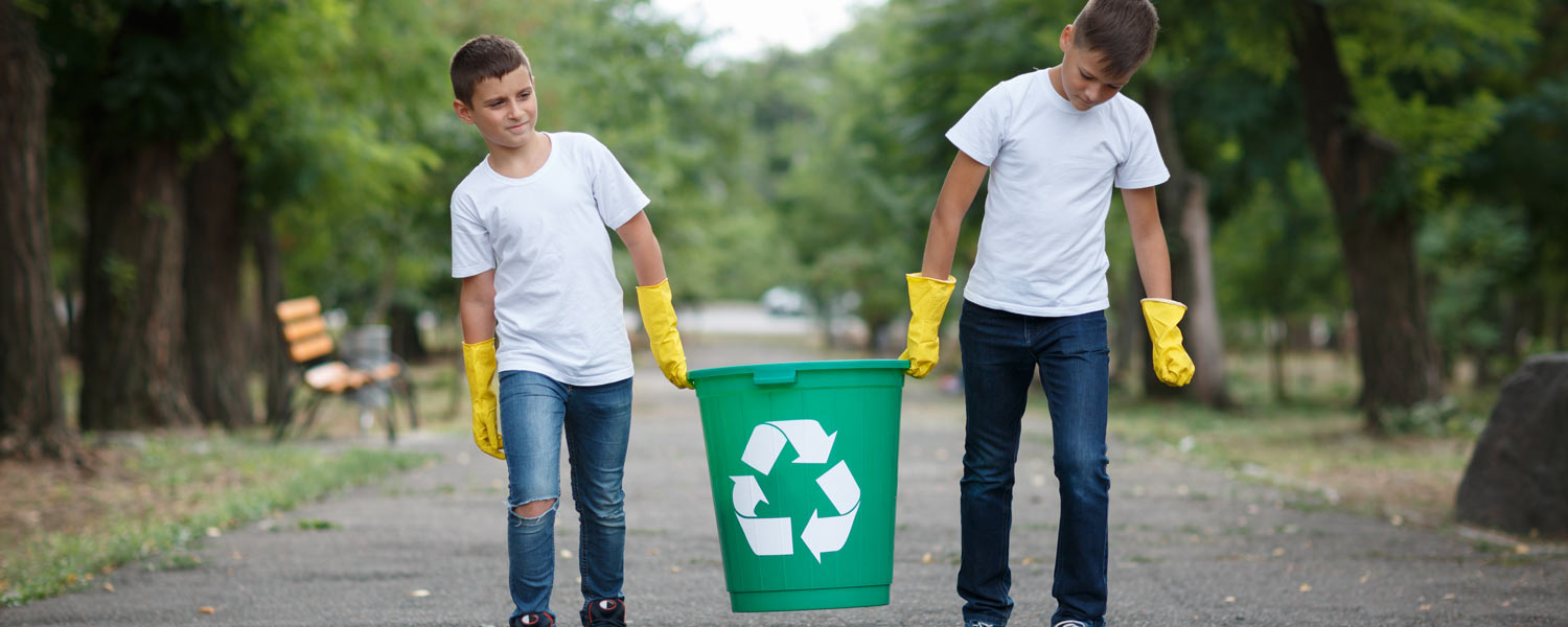 Two boys with yellow cleaning gloves, carrying a recycling bin together