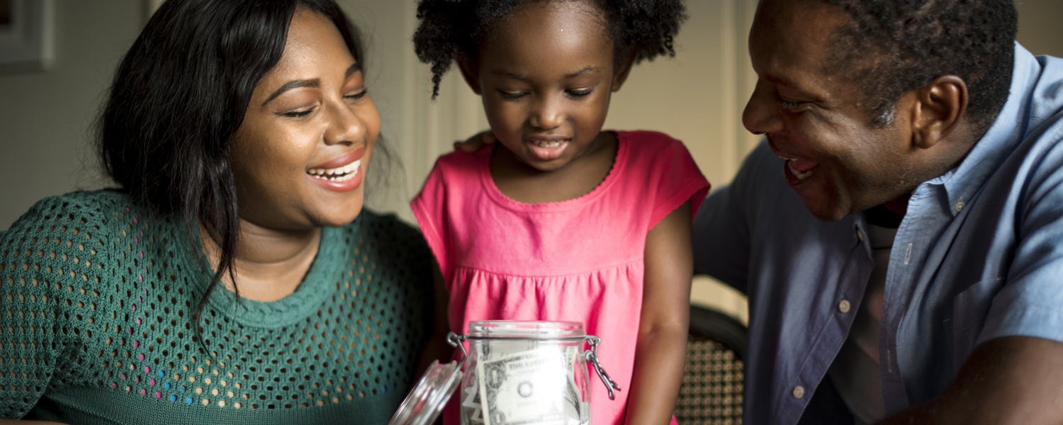 Young girl surrounded by her parents and looking at a jar filled with dollar bills