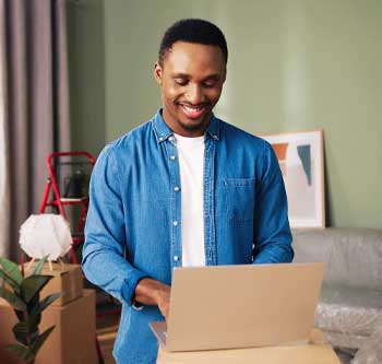 Man standing in the middle of a room surrounded by unpacked boxes, typing on his laptop