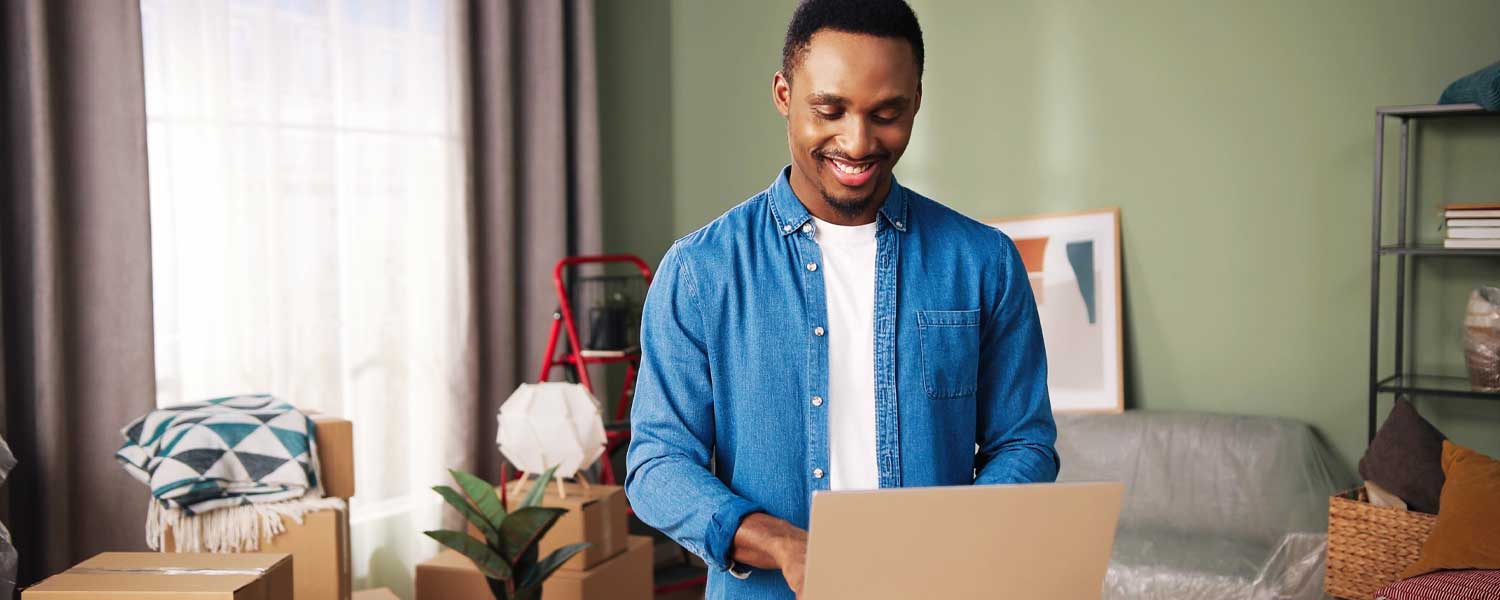 Man standing in the middle of a room surrounded by unpacked boxes, typing on his laptop