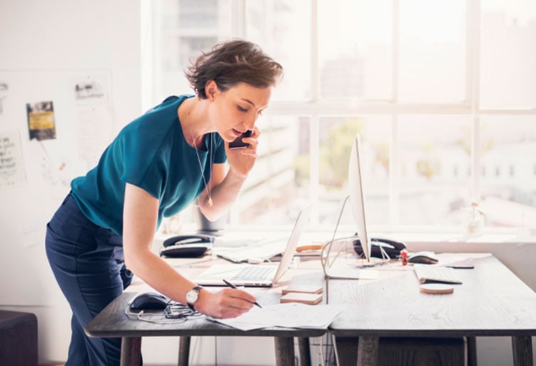 Woman talking on her cell phone while leaning over her desk and writing something down