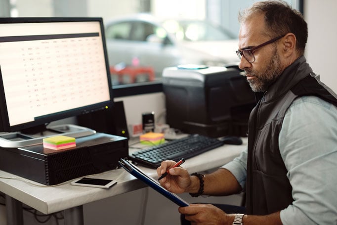 Man writing something in a notebook while financial data is on his computer screen